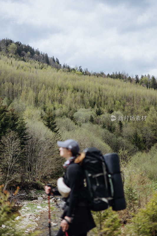 女徒步者背着背包，拄着登山杖站在山上，看着美丽的春景，这是前景的焦点