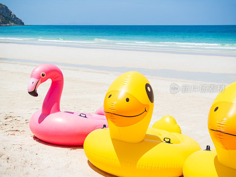 环泳充气浮在沙滩上的海岸与模糊的蓝色大海和天空的背景，游泳池圈黄色橡胶，夏季热带管，粉红色的鸭子火烈鸟床垫在水上，救生圈