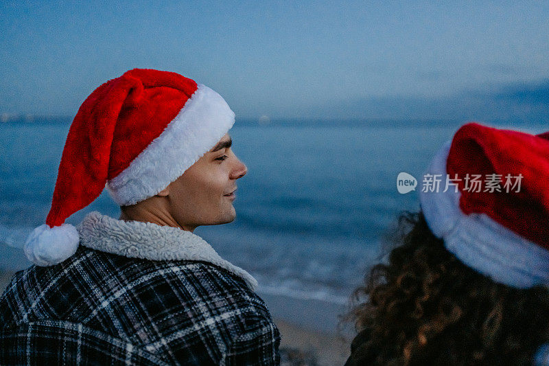 希腊，一对戴着圣诞帽的年轻夫妇在黎明时分欣赏着海岸线的景色