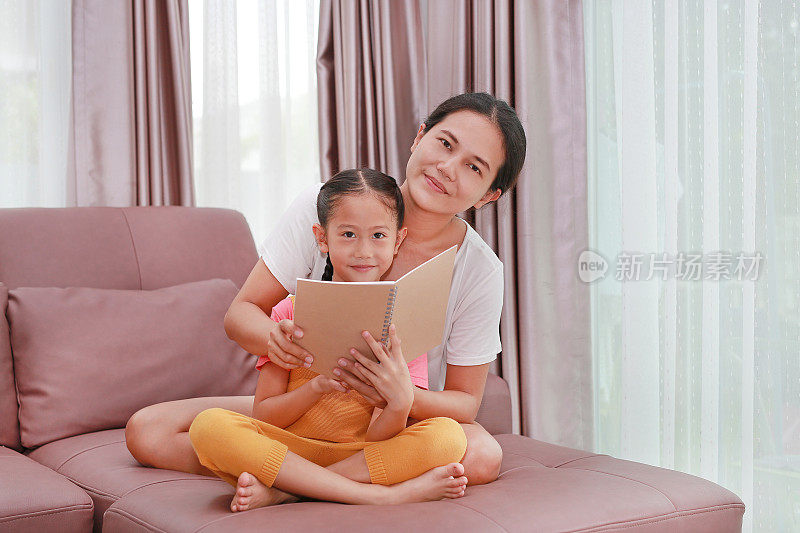 亚洲妈妈和女儿坐在沙发上看书，看着相机。家庭美满幸福