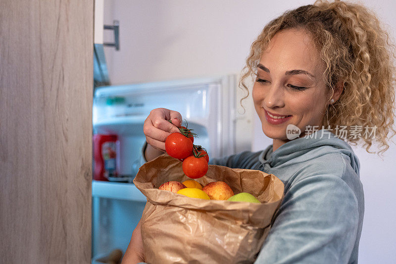一个女孩站在冰箱前，拿着一个装满水果和蔬菜的购物袋
