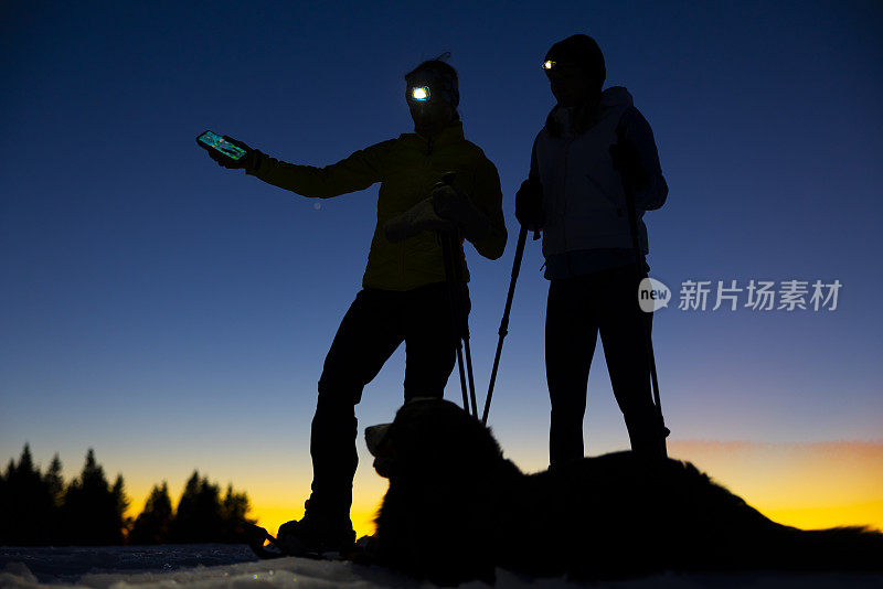夜间雪坡上，两名女滑雪者额头顶着手电筒，站在宠物狗旁边