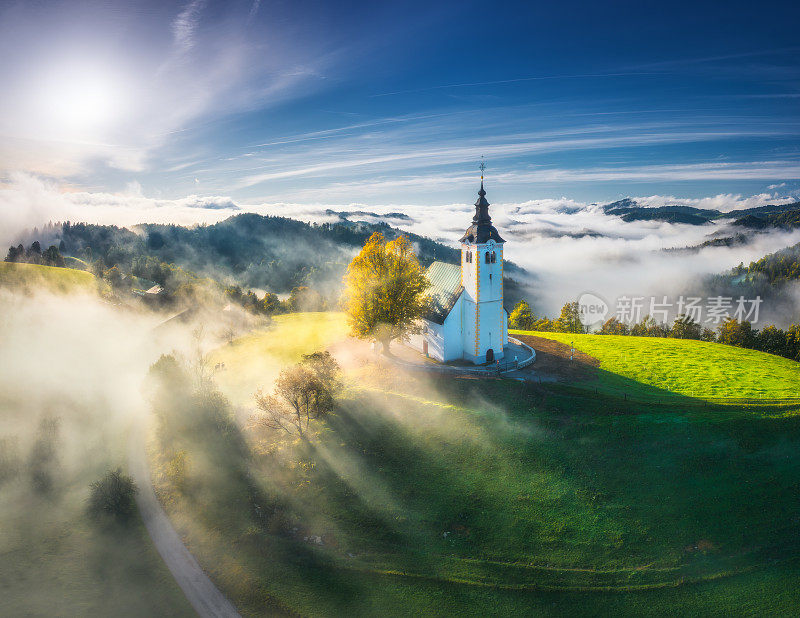 鸟瞰秋天日出时低云笼罩的小教堂。斯洛文尼亚。山顶上美丽的教堂在雾中，绿色的高山草地，树木，蓝天在秋天的黎明。旅行