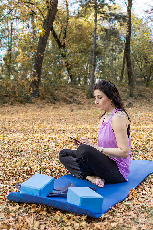秋天，一位中年妇女在树叶环绕的森林里练完瑜伽后正在使用手机。她有锻炼的器材。