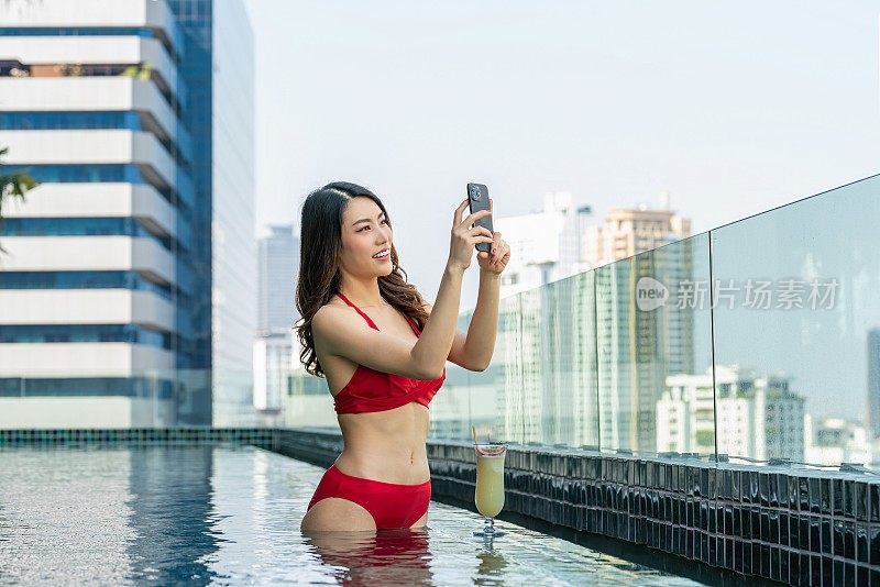 城市景观探索者:站在屋顶游泳池，女人用她的手机捕捉到令人惊叹的景色，在池边喝酒。