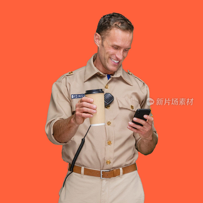 白人年轻男性警察身穿制服站在橙色背景前，手持咖啡杯，使用短信