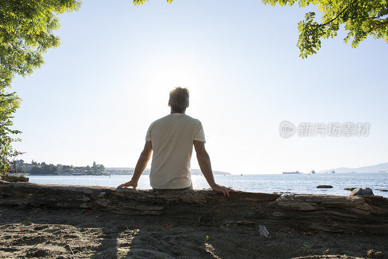 成熟的男人在海滩的原木上休息，海港和大海遥远