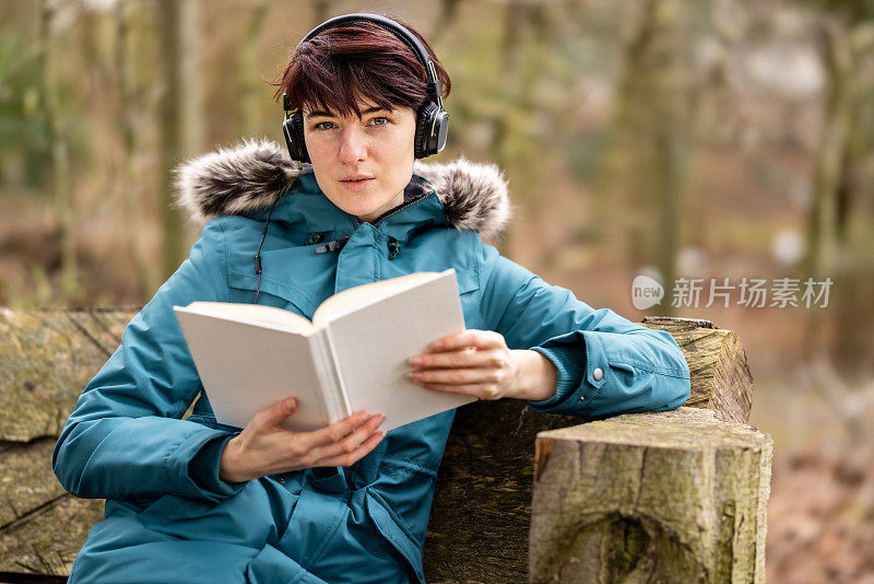 一个穿着蓝色雨衣的年轻女子坐在户外公园的长椅上一边看书，一边戴着耳机听音乐