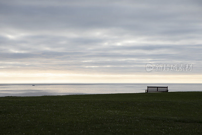 空旷的公共公园长椅上的草地，远处是平静的大海，黎明时分