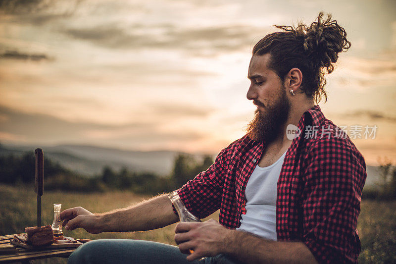 自然的守护者。一个在山上的男人，一个伐木工人，喝着自制的白兰地，吃着自制的食物，而在他身后是美丽的日落，休息辛苦的工作。