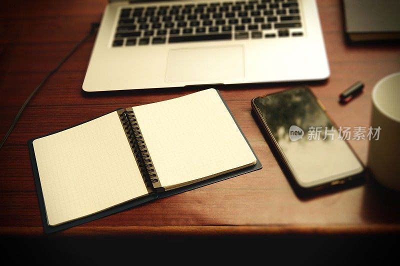从工作台木桌后面拍摄的水平照片，上面有空白页的螺旋形笔记本，笔记本电脑，手机，咖啡杯