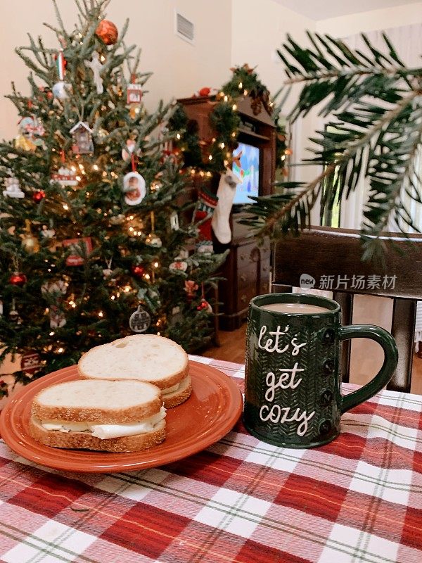 温馨的圣诞早餐:两个煎蛋三明治，热咖啡放在写着“让我们变得舒适”的圣诞杯里，站在圣诞树前，在家过一个温馨的圣诞