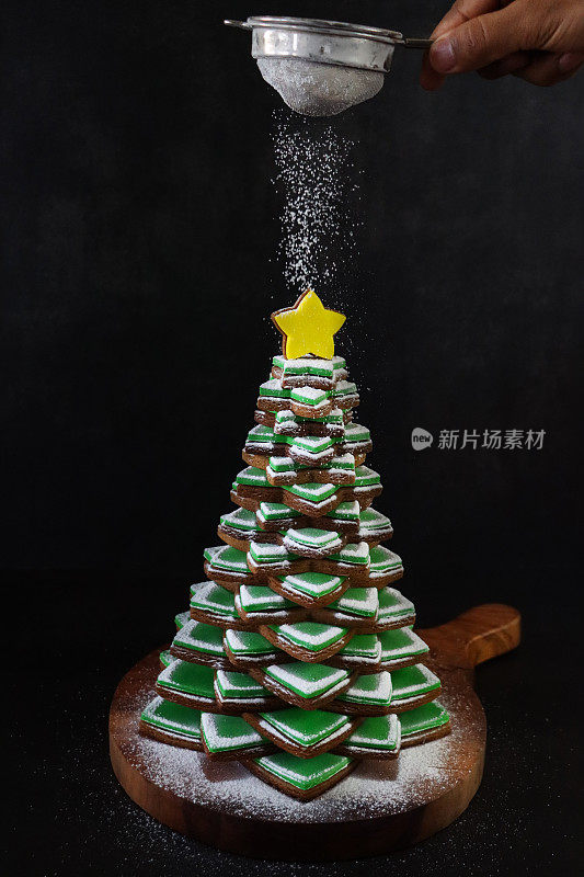 难以辨认的人在圣诞树上用筛子筛糖粉的形象，上面堆叠着绿色方糖糖霜的姜饼星饼干，黄色冰镇星饼干顶部，木制蛋糕架，黑色背景