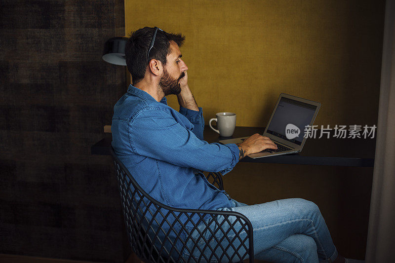 一个留着胡子的嬉皮士坐在桌子旁工作。他正在使用笔记本电脑。在他的笔记本电脑旁边有一个咖啡杯。