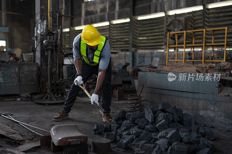 非洲裔美国男性工人穿着背心和头盔安全使用铁锹在金属工厂的地面上挖煤燃烧。在工业工厂操作重金属机械的技工领班。