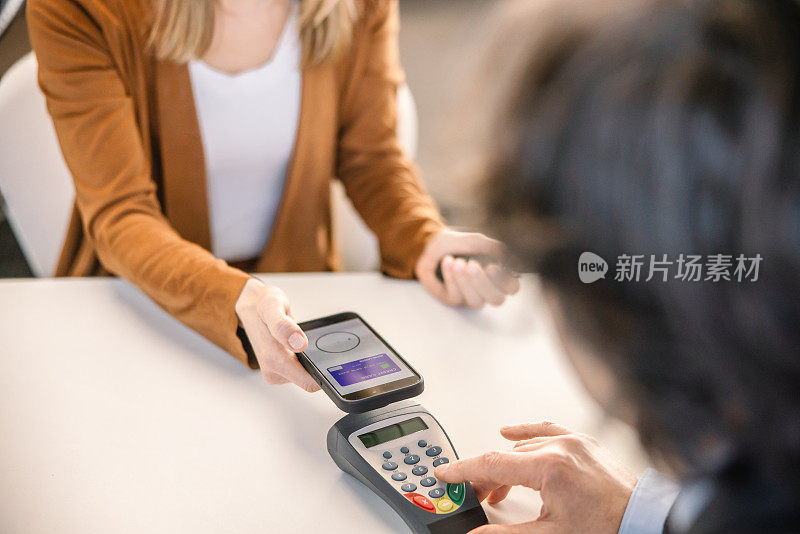 一名女顾客正在汽车销售店的室内办公桌旁，与销售人员一起，用NFC(近距离通信)技术用智能手机支付新车费用