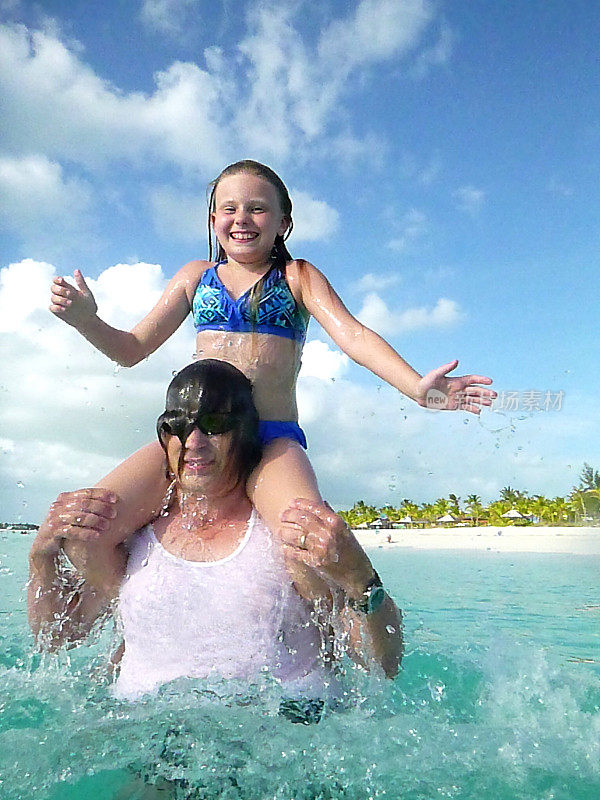 一名50多岁的成年白人男子戴着太阳镜、手表、白色t恤和泳裤，在一个阳光明媚的日子里把坐在他肩膀上的10岁女儿扔进海里，特克斯和凯科斯群岛
