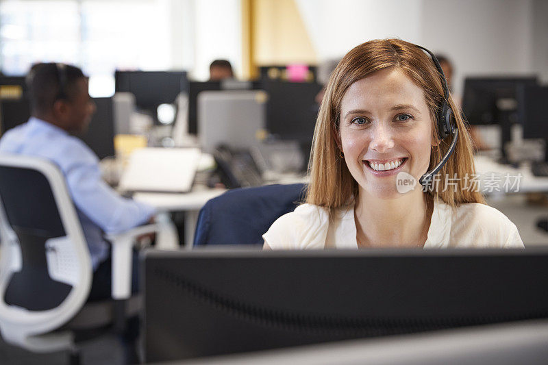 在繁忙的办公室里戴着耳机在电脑前工作的年轻女子
