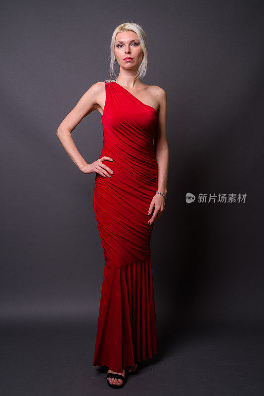 漂亮的金发女人穿着红色的不对称长裙对抗灰色的背景