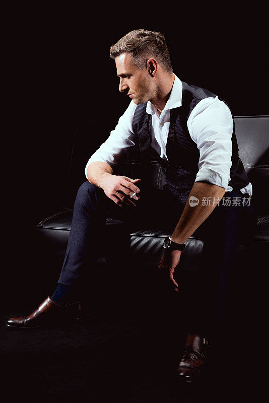英俊的男子穿着正式的服装坐在沙发上，用香烟孤立在黑色