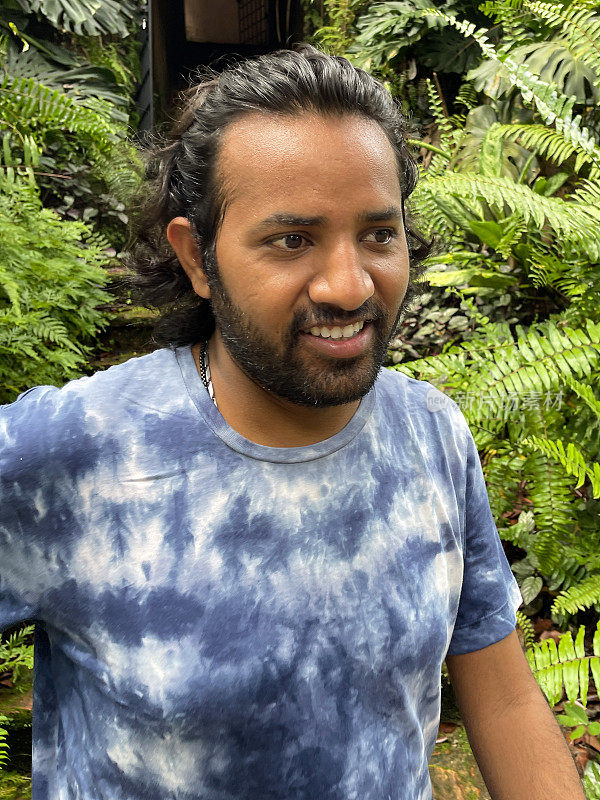 印度男子穿着休闲服在热带花园散步的特写图像，穿着自制的扎染t恤，蕨类植物，重点在前景