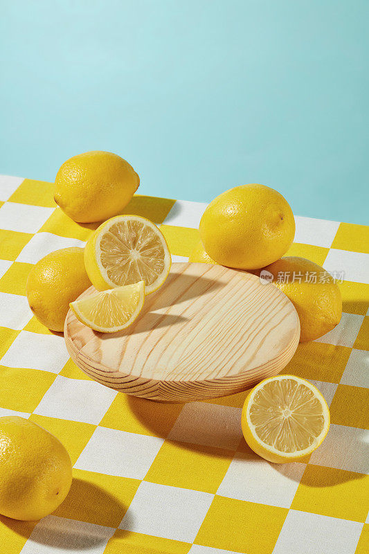 柠檬主题的垂直照片，黄色格子平面与一个空白的圆形木制底座和充足的新鲜黄色柠檬在上面，在淡蓝色的背景。展示产品留白，高角度