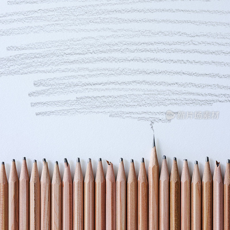 一排钝的，磨损的木制绘画铅笔在白色的画纸背景上，一个木制铅笔是非常尖锐的从人群中脱颖而出，与钝的铅笔形成对比，在纸上的铅笔画阴影区上方。