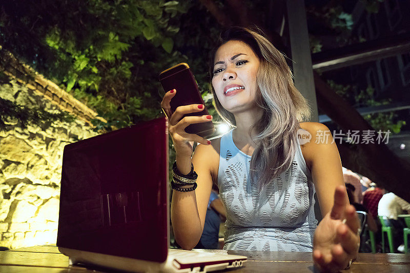 一个愤怒的女人查看手机，在酒吧里使用笔记本电脑