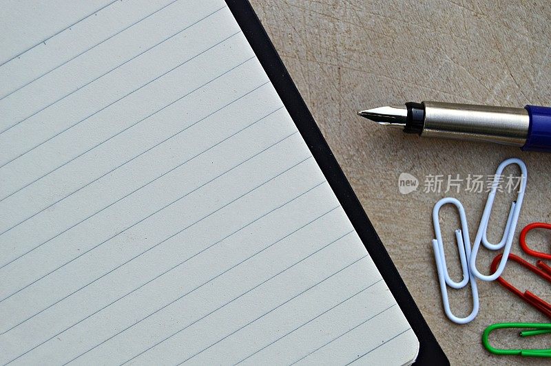 钢笔、回形针和日记——一张经典的水平照片，一张空白的白色单行页的记事本，黑色封面，蓝色和银色的钢笔和彩色回形针放在一个木制的米色旧古董