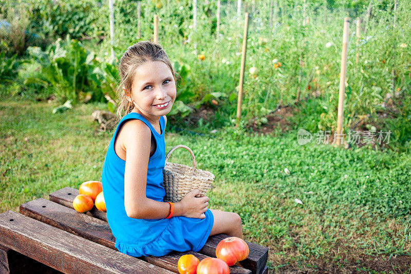 可爱的微笑女孩拿着篮子坐在蔬菜园里