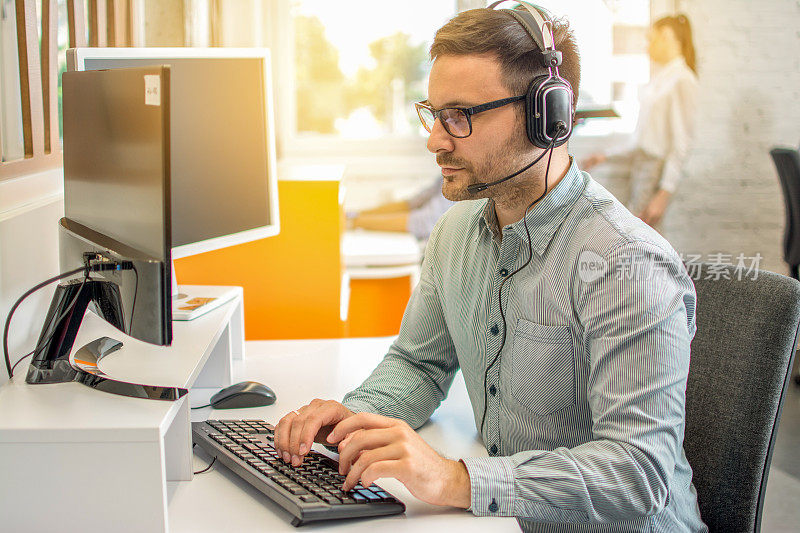 男性专业呼叫中心电话销售代表戴耳机使用电脑在客户服务支持服务办公室。