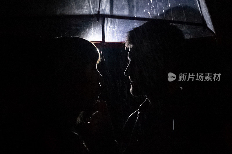 一对相爱的情侣在伞下接吻，而雨却落在他们身上。人的夜间摄影。保护夫妻关系和长久的爱的概念。