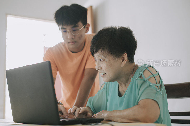 在餐厅里，一位年长的亚洲华人女士向她的孙子学习如何使用笔记本电脑社交媒体