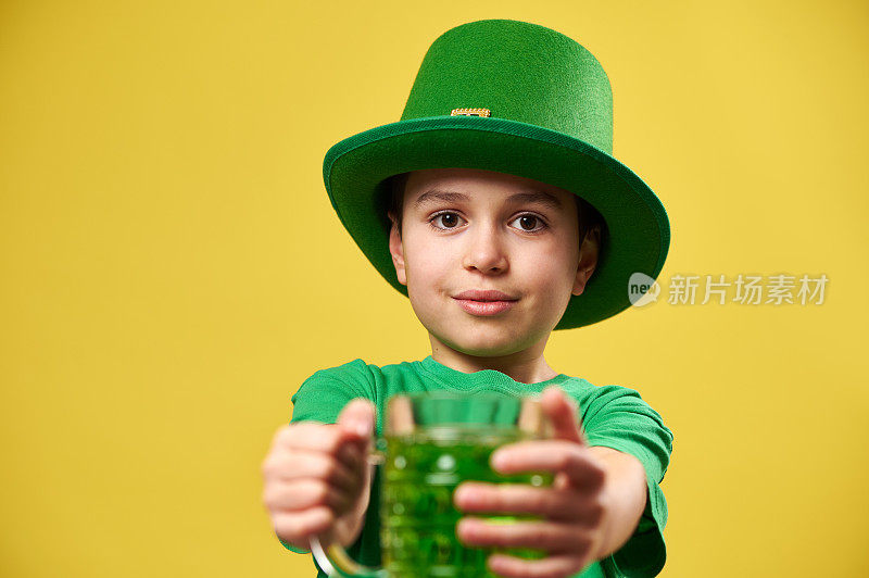 一个戴着矮妖精帽子的男孩拿着一杯绿色饮料站在镜头前。孤立在黄色背景上。圣帕特里克日
