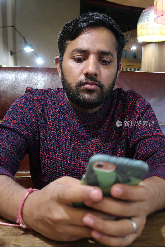 一名印度男子坐在餐厅的餐桌前等待点菜，一边看手机短信