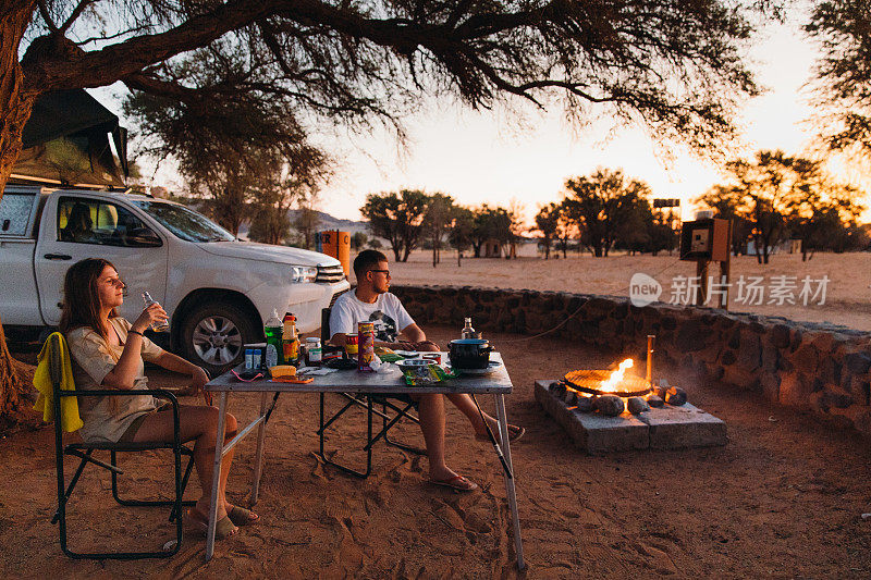 在纳米比亚的沙漠里，夕阳西下，男女旅行者开着露营车在树下露营