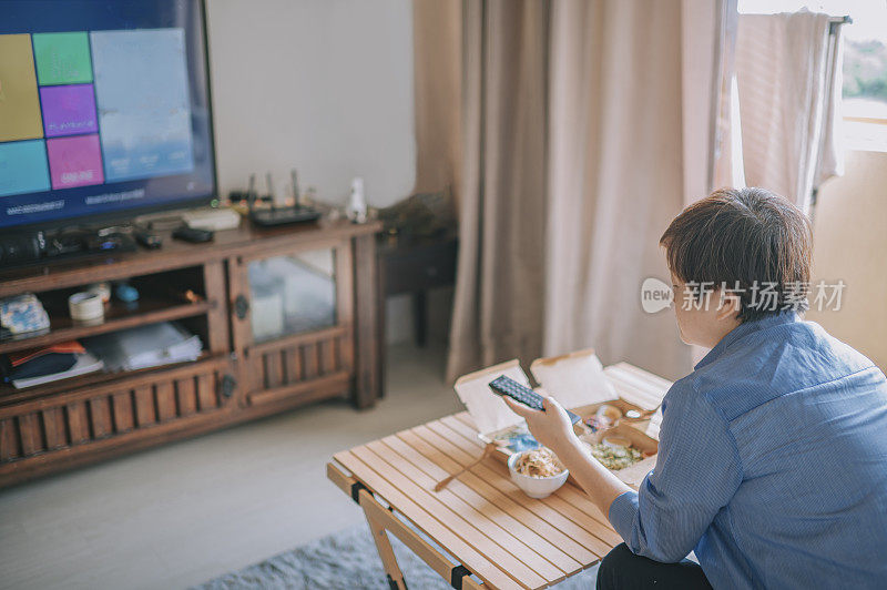 一名亚洲华人女子在客厅里用再生纸容器一边使用电视遥控器一边选择频道，一边吃着外卖，包括马来西亚炒饭、乌兰炒饭和印度炒饭