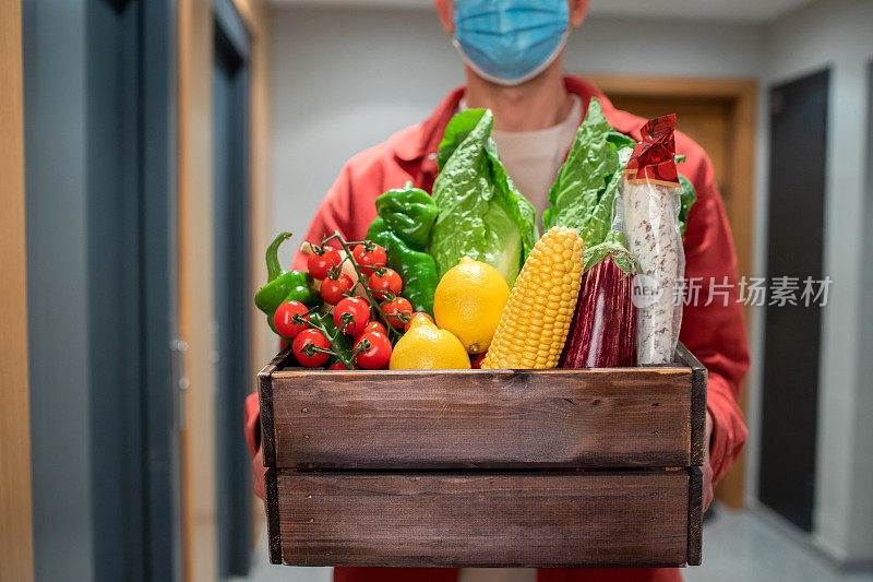 带防护口罩的送餐员手持纸袋，携带食物进入入口。快递员把装有新鲜蔬菜和水果的盒子交给顾客
