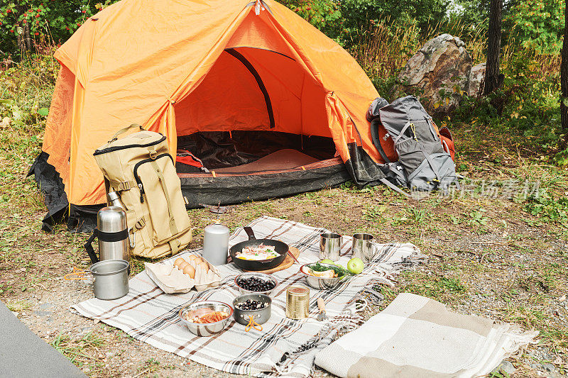 在森林里，在橙色帐篷和背包的旁边，在炉子上烤的各种菜肴被放在布上