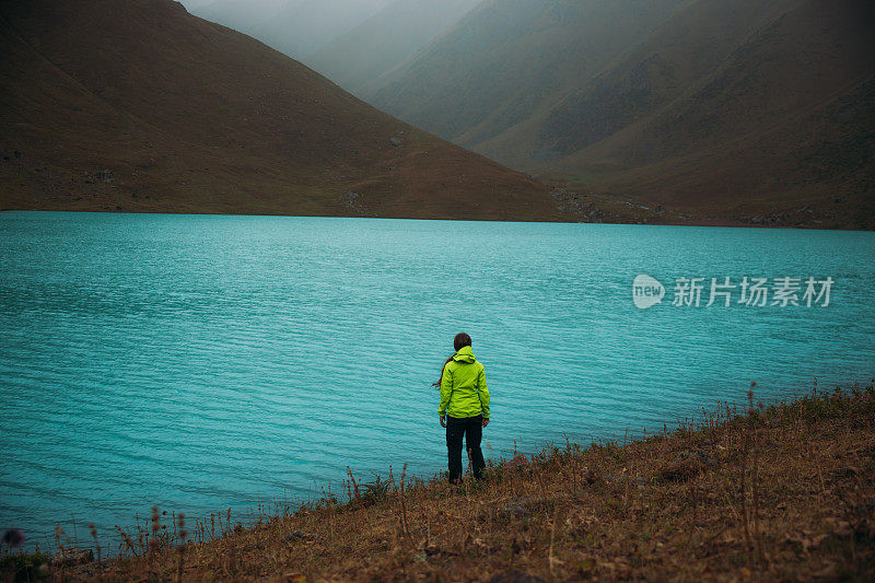 一名女子背包客坐在中亚绿松石山湖畔，凝视着大自然