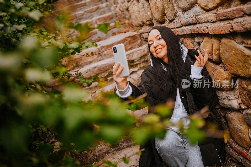 年轻快乐的女游客独自旅行，用智能手机与景点自拍，视频聊天。Blogger直播流。深色头发的女士穿着黑色羽绒服和白色连帽衫