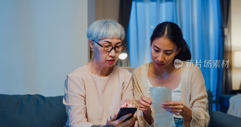 幸福的亚洲家庭，年幼的女儿和年迈的妈妈坐在沙发上，晚上在家里教年迈的妈妈如何使用智能手机支付在线支付设备账单。