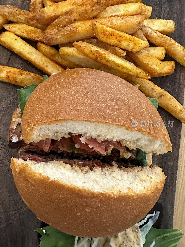 汉堡的特写图像，培根，番茄，泡菜，生菜和蛋黄酱在无籽汉堡面包切成两半在木制砧板上与脆脆的薯条，木纹背景，高视图