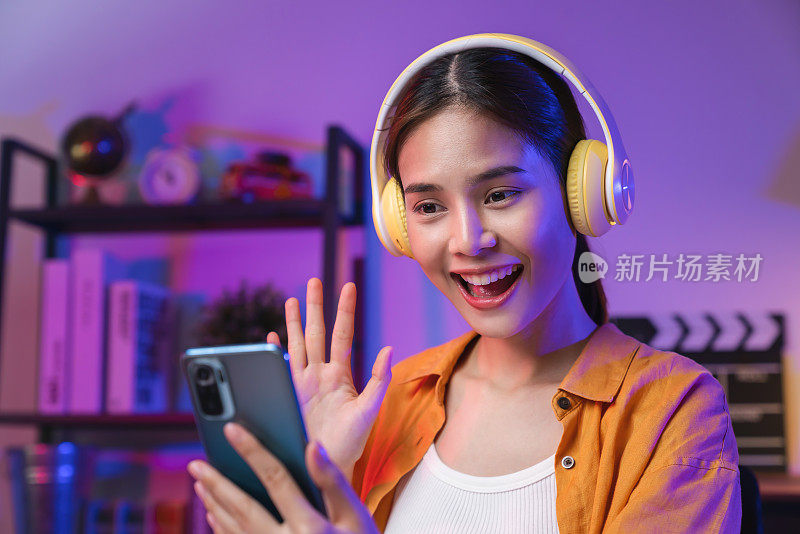 开朗美丽的亚洲女人戴着耳机，看着智能手机在网上视频通话会议。