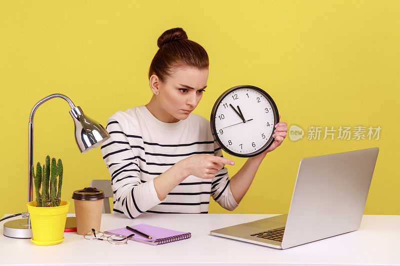 女子手持大挂钟，管理着时间，用严肃的表情指着笔记本电脑屏幕。