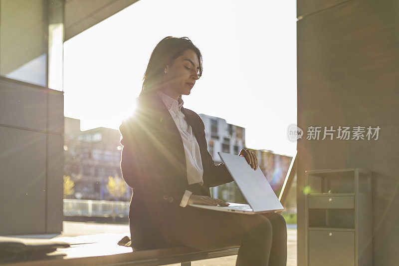 一个穿着优雅西装的女孩在户外辛苦工作后，坐在公司大院里的长凳上合上笔记本电脑。