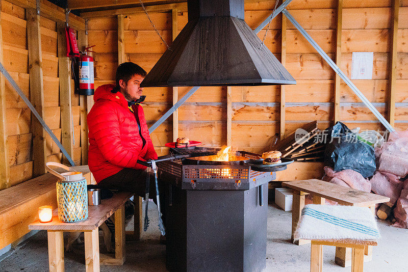 在挪威，一名身穿红色夹克的男子在真正的木制掩体里用火烹饪肉