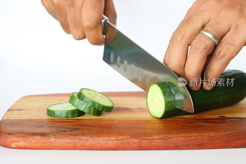 不可识别的人用菜刀在木制砧板上切黄瓜片的特写图像，整个水果和切片显示绿色的皮，白色的肉和白色的种子，白色的背景，重点放在前景上