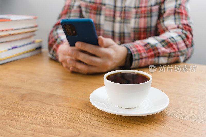 木桌上的一杯咖啡，一个面目全非的人在用智能手机，坐在桌前，手里拿着一摞书，聚焦在前景