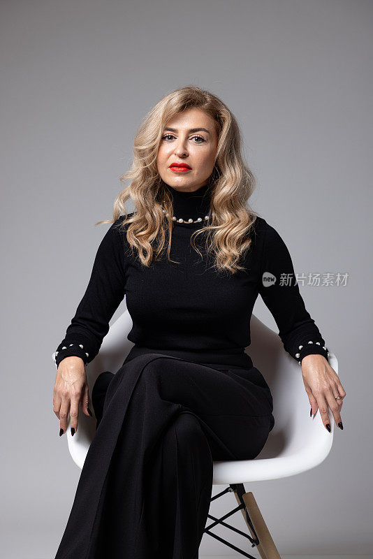 穿着黑色连体衣的时尚女子坐在白色的椅子上，摆出微妙而自信的姿势。